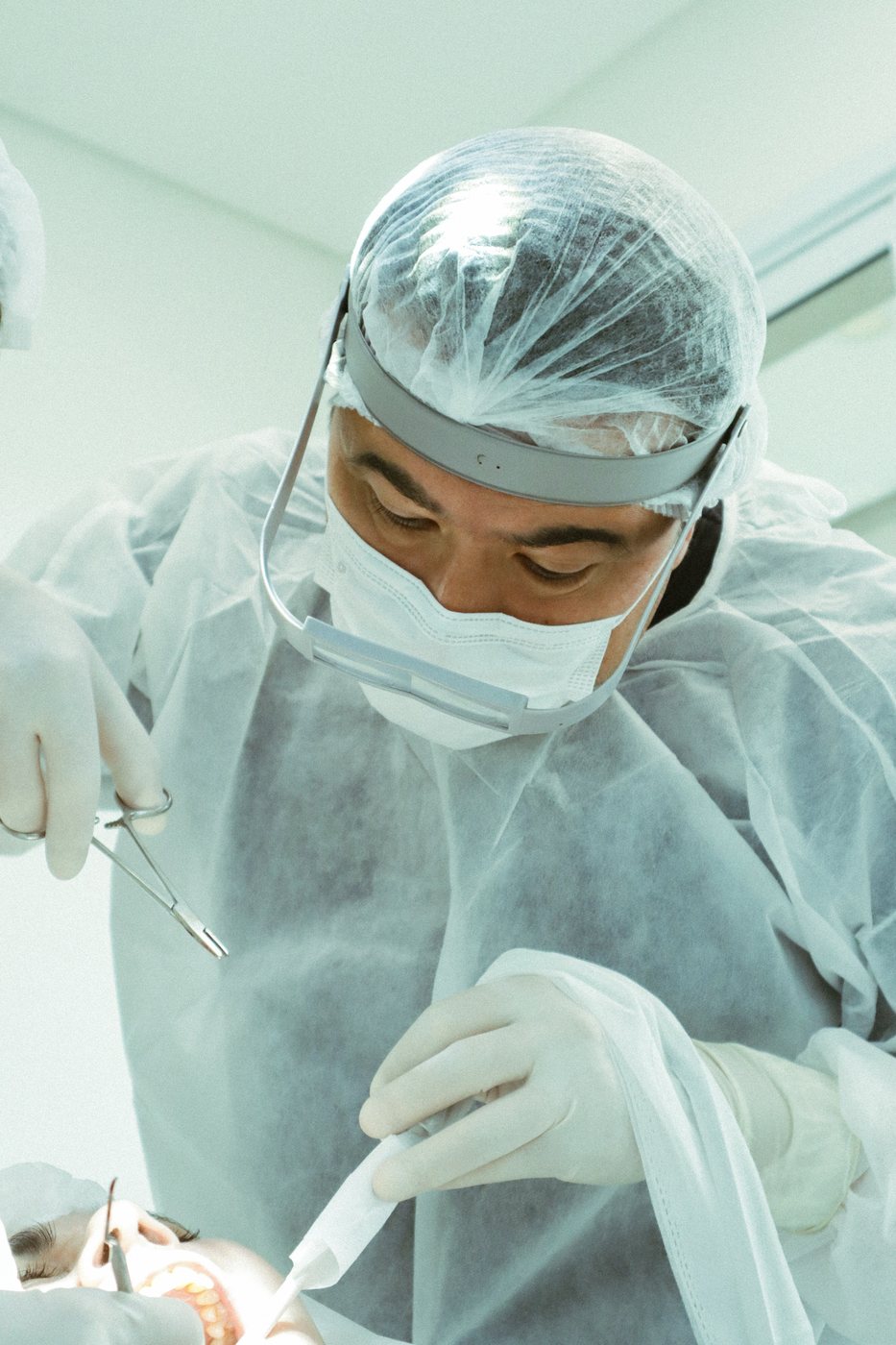 Cirurgião-dentista de touca, máscara e protetor facial operando, visto de baixo em luz clara